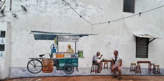 Malakka’da bir sokak duvarında yer alan seyyar satıcı temalı duvar resmi önünde oturan bir ziyaretçi