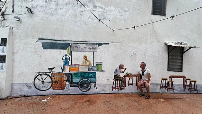 Malakka / Malezya Malakka’da bir sokak duvarında yer alan seyyar satıcı temalı duvar resmi önünde oturan bir ziyaretçi